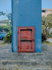 Naklejka premium A vibrant blue wall features a weathered red door at its base, surrounded by greenery and pavement, creating an intriguing urban scene.