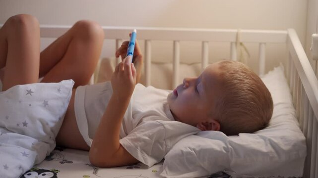 Child playing on tablet lying in bed in evening. Cute little boy watching cartoons on mobile before go to sleep. Boy enjoying in bedroom looking on screen.