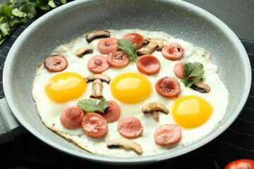 Tasty fried eggs with cut sausages and mushrooms in frying pan on black table, closeup