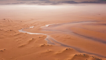Desert Oasis: Aerial view of a winding waterway cutting through the vast expanse of the sandy desert, creating a striking contrast of textures and colors under a hazy sky. 