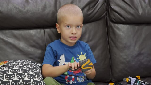 Child playing with new toy sitting on sofa at home. Concentrated face of little boy holding transformer robot toy. Three year old child examining new toy comments on his actions.