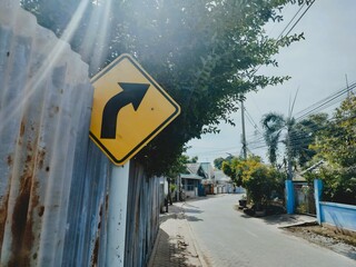 A yellow right turn road sign is visible along a narrow street lined with greenery and fences under bright daylight.