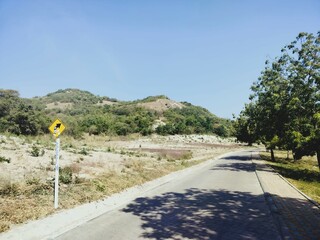 A serene road lined with trees, leading towards rolling hills under a clear blue sky, showcasing a...