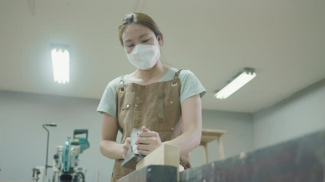 A woman wearing a mask diligently carves wood in a well-lit workshop, showcasing craftsmanship and focus. Her workspace is filled with tools, emphasizing the art of woodworking and creativity in actio
