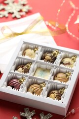 Box with tasty chocolate candies in shape of baubles and Christmas decor on red background, closeup