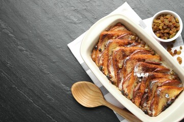 Freshly baked bread pudding in baking dish, raisins and wooden spoon on dark textured table, top view. Space for text