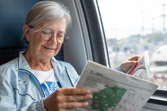 Senior smiling woman with eyeglasses enjoys the train ride sitting in passenger seat by the window reading a newspaper - Powered by Adobe