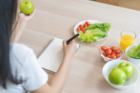 Diet, happy asian young woman working, write diet plan right nutrition holding green apple with eat fresh vegetable salad is food for good health. Nutritionist for weight loss of healthy at home.