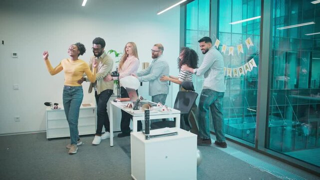 Working team moving in line like train while celebrating birthday their coworker. People laughing and joking during walking around table. Room decorated with colorful banner. Warm and joyful vibe.