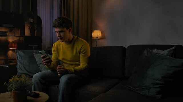 Young man in the evening after work relaxing at home, sits on the sofa in living room watching tv, holds smartphone anticipating getting good news, winning expression.