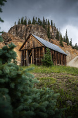Abandoned wooden mine building nestled in a scenic mountain setting with dramatic rocky cliff and evergreen trees, evoking rustic charm and natural beauty.
