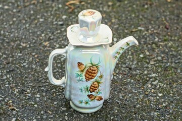 one beautiful white ceramic kitchen teapot with handle and closed round lid for brewing hot tea with a pattern small teapot stands on black asphalt during the day outside