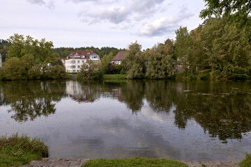 Obraz premium Serene River Landscape with Cozy Cottages Nestled in Lush European Greenery Under Dramatic Twilight Sky. Tranquil European Riverside Village at Dusk: Picturesque Houses Nestled Among Lush Greenery