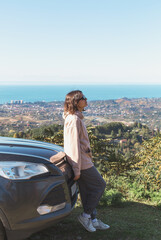 Fototapeta premium A young woman in casual clothes leaning against a car, enjoying a scenic view of the sea and the city below from a hilltop, surrounded by greenery under a clear blue sky, concept of travel and freedom