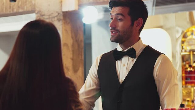 Good looking smiling bartender friendly recommending special drink list for VIP customer at beverage counter bar luxury, making great first impression in neat professional barman costume. Vinosity.