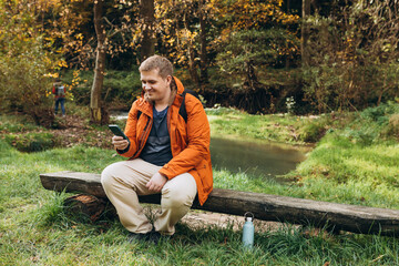 Stylish man with backpack holding smart phone and relaxing in nature. Full body photo. Hiking active male lifestyle siting on tree trunk outdoors. High quality photo. Adventures.