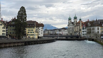 A serene view of Lucerne with historic buildings, a baroque church with green domes, and snow-capped mountains in the distance under a cloudy sky, creating a charming and picturesque scene.