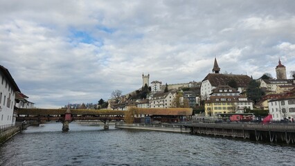 A scenic view of Lucerne with a wooden covered bridge spanning the water, historic buildings, towers, and a hilltop castle under a cloudy sky, creating a picturesque European landscape.