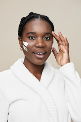 A joyful young woman of African descent applying skincare with a smile, wearing a cozy white robe,...