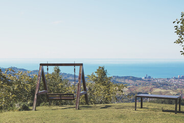 Wooden swing and bench on a grassy hilltop overlooking the sea and a distant city under a clear blue sky, evoking peace and simplicity