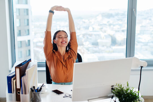 Happy lady manager sitting on chair and stretching her back and arms, taking break, feeling pleased and satisfied at workplace in office