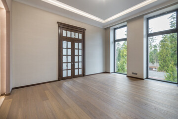 A spacious empty room with the final finish in a luxurious mansion. Brown door with frosted glass windows. Panoramic windows overlooking the green landscape. Unfurnished.