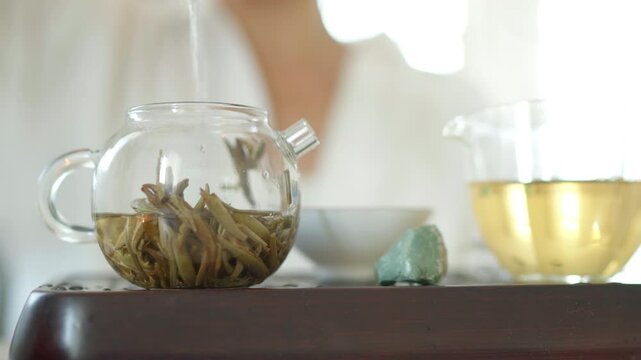 Female pouring hot water in clear glass teapot with aged single bud creating serene ritual. Soft light, pitcher on wooden tea tray evoke calming mindful Chenese ceremony atmosphere 4K video