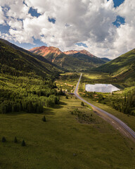A breathtaking view of a green valley framed by mountains, featuring a calm lake and a winding road. The sunny sky and scattered clouds enhance the beauty of this tranquil landscape.