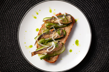 A gourmet open sandwich with sardines on whole grain bread garnished with cucumber slices and green herbs on a white plate