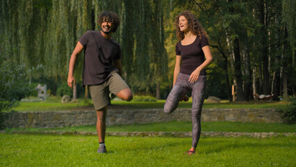 Active healthy couple Indian Arabian man and Caucasian woman athletes doing sport morning workout stretching body muscles warm-up exercises in park outdoors sporty people diverse male female training
