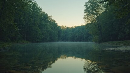 Obraz premium Misty sunrise over a calm lake surrounded by lush green trees.