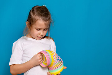 Fototapeta premium Beautiful happy blonde little girl plays with a plastic colored neon rainbow spiral children toy, holding bouncy stretchy toy. 90s toy, nostalgia, blue background, copy space