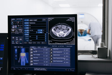 In the MRI or CT control room with a monitor displaying brain scan results, while a patient is being prepared for the examination in the background. The concept of precise diagnostics and technology.