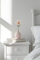 Pink flower in a vase on a white bedside table, next to a bed with white bedding. Sunlight streams through a window.