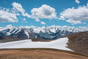 Aerial top view to stony pass and snow-white glacier on rocky hill in sunlight among high mountains in sunny day. Large colorful mountain range with snowy peaks in far away under clouds in blue sky.