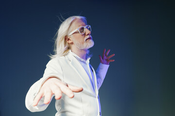 Portrait of a man with long white hair and glasses, dressed in a white suit, striking a dynamic...