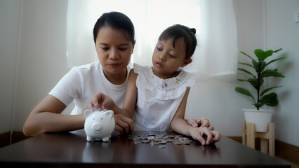 A woman and a child are sitting at a table with a piggy bank. The woman is putting coins into the piggy bank while the child watches. Concept of financial responsibility