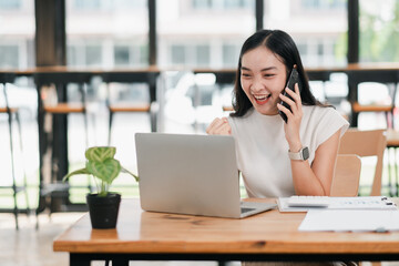 Excited young woman using a laptop and smartphone in a cafe, celebrating success. Modern workspace with natural light.