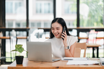Obraz premium Smiling woman multitasking with a smartphone and laptop in a bright, modern office setting.