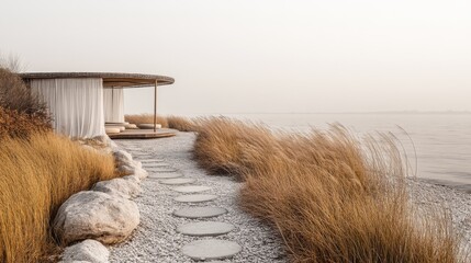 A quiet walkway paved with smooth stones, surrounded by tall grass, leads to a private beach pavilion. The pavilion has airy white curtains and beige tones, creating a peaceful, fresh and clean feel.