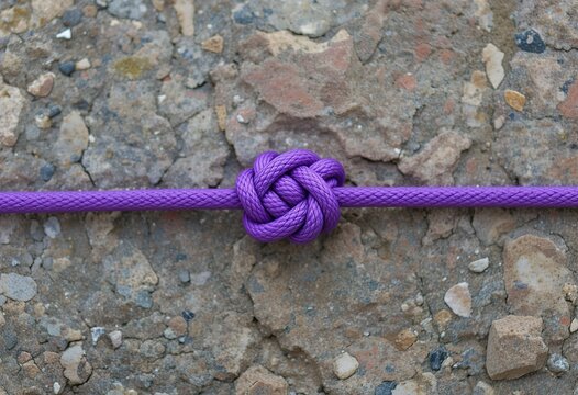 A close-up photograph shows a single purple knot tied in a rope against a rough, textured stone background.