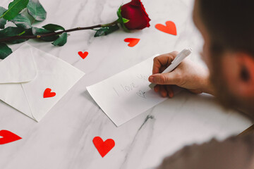 A man writes a heartfelt note saying I love you on an envelope, surrounded by red hearts and a single rose on a marble table, creating a romantic atmosphere. Romantic gesture. Happy Valentine's Day.