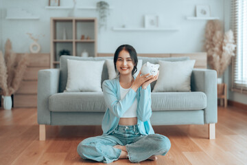 Young Asian woman holding piggy bank and sitting on the floor at home. Save money and financial investment