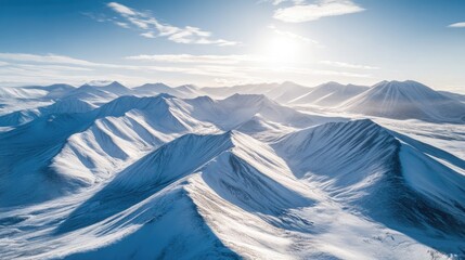 Aerial shot of vast snowy mountains, sun-drenched peaks casting long shadows, with clear skies and a few wisps of clouds hovering above the landscape.