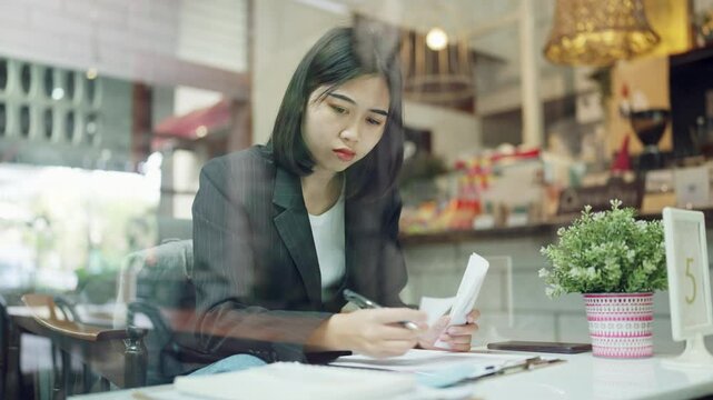 A woman in a business suit is sitting at a table with a stack of papers and a receipt in front of her. She is focused on the paperwork, possibly reviewing or organizing it