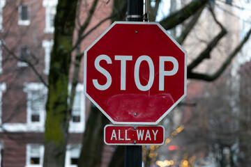 A red octagonal stop sign with an "All Way" label below it, positioned on a city street. Surrounded by bare winter trees and urban buildings in the background, capturing a quiet city moment