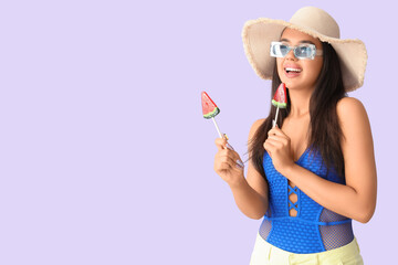 Beautiful happy young woman in beachwear and with watermelon lollipops on lilac background