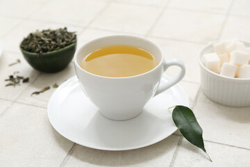 Cup of hot green tea with sugar cubes and dried leaves on white tile background, closeup