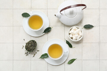 Cups of hot green tea with sugar cubes and dried leaves on white tile background