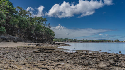 Low tide in the ocean. Boulders are scattered on the exposed seabed. Green vegetation on a coastal cliff. Boats and buildings on the shore are visible in the distance. Blue sky, clouds. Madagascar.